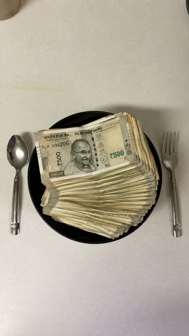 A stack of Indian ₹500 rupee notes (including one with serial number 7LR 934206) sits on a black plate flanked by a spoon and fork, visually symbolizing financial success "as your daily goal" through trading.