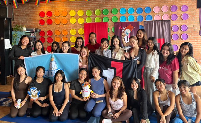 The image features a group of women posing together, possibly at an event or gathering. They are all dressed in black clothing, with some wearing tank tops as well. The women appear to be enjoying themselves and are likely friends or acquaintances. In the background, there is a wall adorned with colorful plates hanging on it, adding a vibrant touch to the scene. A banner can also be seen, possibly indicating that this event is related to blockchain technology or has a connection to the Philippines Blockchain Week. The image captures a moment of camaraderie and celebration among these women.