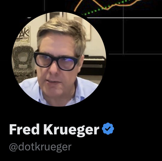 A social media profile image of Fred Krueger (@dotkrueger) featuring a man wearing thick black glasses and a light blue shirt.
