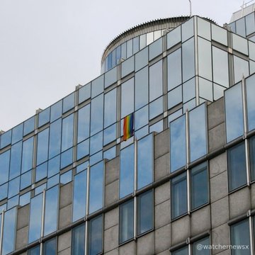 A low-angle exterior shot of a modern glass office building with a rainbow pride flag hanging from a window, credited to '@watchemewsx'.