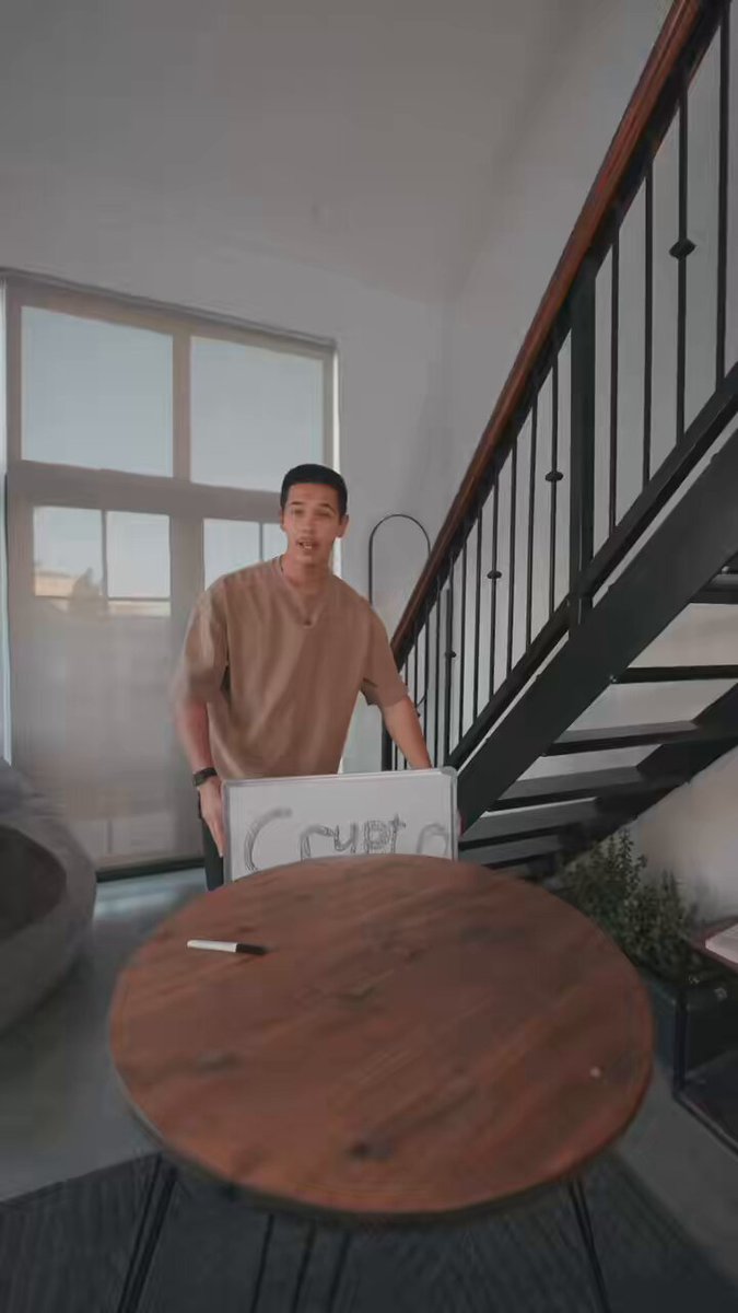 The image shows a man standing next to a wooden table with a white sign that says "Crypto" on it, possibly indicating an upcoming launch of a decentralised trading product or cryptocurrency exchange. The man appears to be presenting something related to this project, as he is holding up the sign and talking about it.
