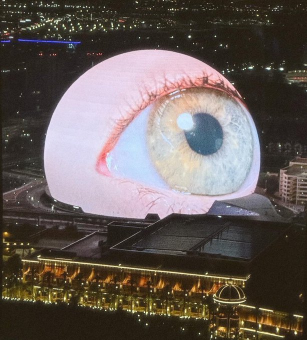 The MSG Sphere in Las Vegas displays a massive, lifelike image of a human eye on its exterior LED screen at night, with the surrounding cityscape illuminated in the background.