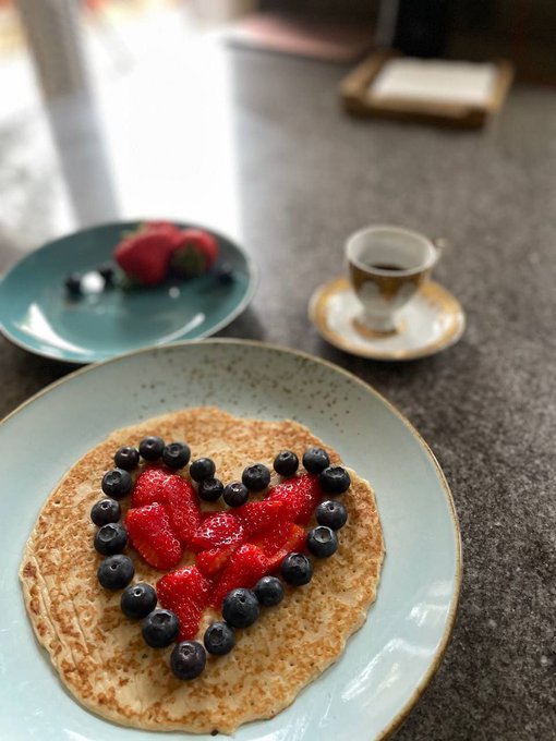 A light blue plate holds a crepe topped with fresh strawberries and blueberries arranged in a heart shape, accompanied by a side plate with more strawberries and a small cup of espresso.