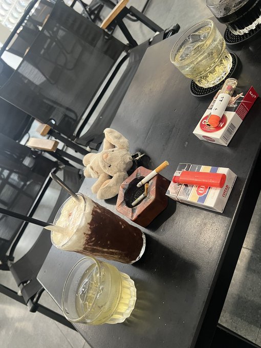 An overhead view of a cafe table holding two glasses of yellow drinks, an ashtray with cigarettes, a small plush toy, and a pack of Marlboro cigarettes.