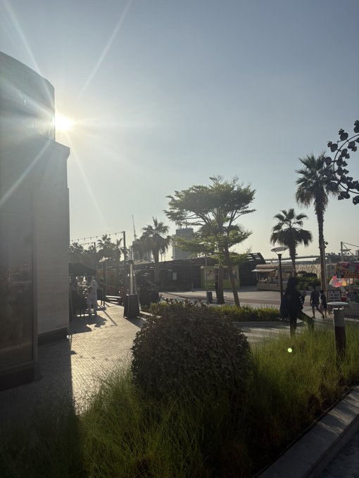 A sunny outdoor scene at what appears to be a GM-branded event space, featuring palm trees, landscaped gardens, and people walking near pavilion structures with the sun casting a bright glare over the venue.