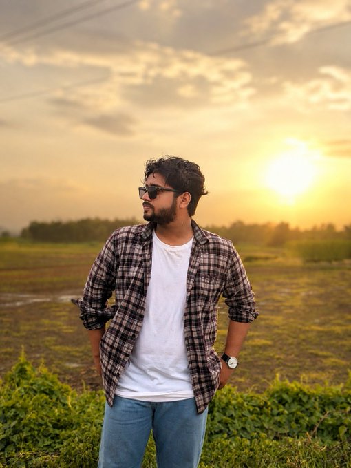 A young man wearing sunglasses, a white t-shirt, and a plaid shirt stands outdoors during a golden sunset in a grassy field.