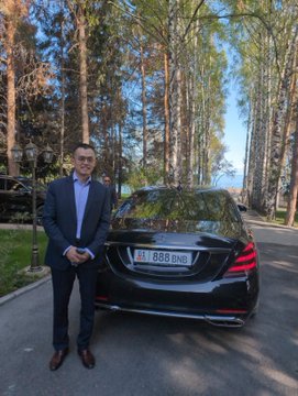 A man in a blue blazer and sunglasses stands smiling next to a black Mercedes-Benz sedan with license plate "AA 888 BNO" in a wooded area.