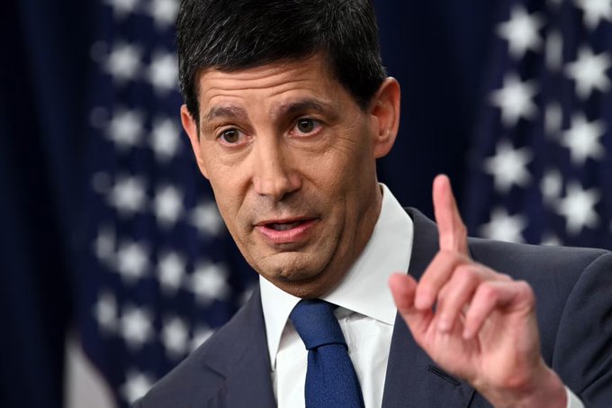 A close-up photograph of a man in a dark suit and blue tie speaking while pointing his finger upward, with American flags blurred in the background.