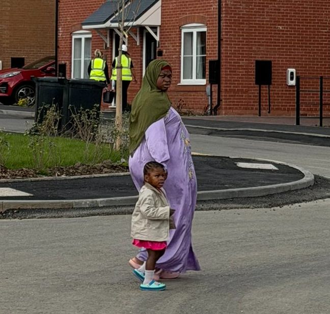 A woman wearing a green hijab and purple traditional dress walking with a young child in a pink skirt and beige jacket on a street in a residential area with brick houses.
