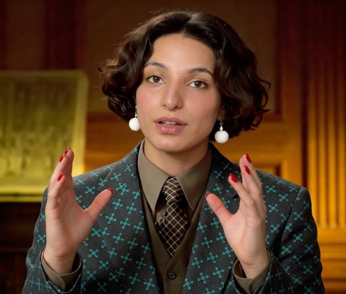 A woman with short dark hair, wearing a patterned teal and gray blazer over a brown shirt and plaid tie, gestures expressively with both hands while speaking, accessorized with white pom-pom earrings and red nail polish.