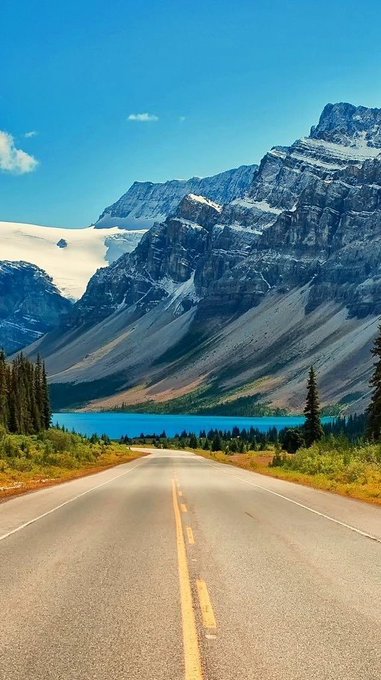 A scenic view of a road leading toward a turquoise lake surrounded by majestic snow-capped mountains under a clear blue sky.