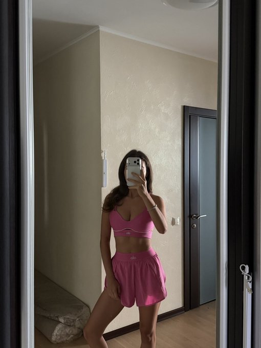 A mirror selfie shows a woman in a pink Alo sports bra and matching pink athletic shorts taking a photo with her smartphone in a room with a closed grey door.