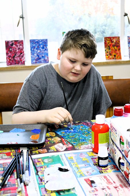 A young boy in a gray shirt paints at a table with various art supplies, including brushes, paint bottles, and colorful artwork in the background.