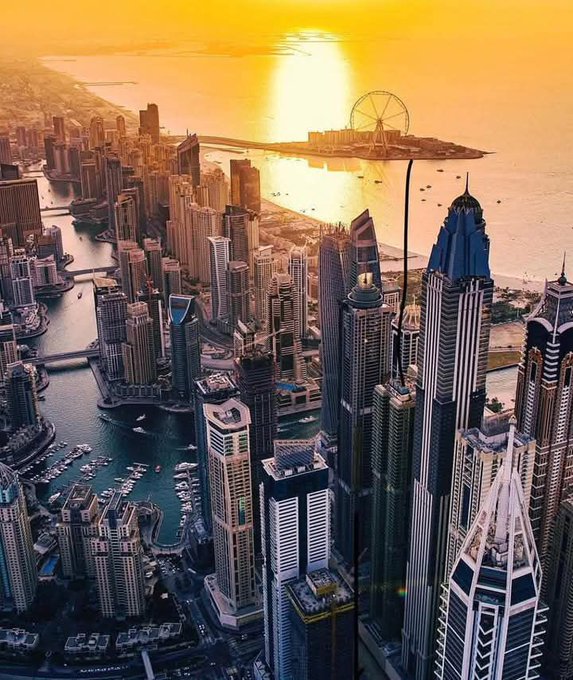 Aerial view of the Dubai Marina skyline at golden sunset, featuring a surreal floating platform with a Ferris wheel hovering above the city.