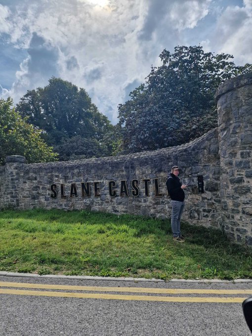 A man in a black hoodie and cap stands beside a stone wall engraved with 'SLANE CASTLE' under a partly cloudy sky.