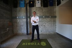 A man standing with arms crossed in an industrial space with a green 'LIBER8' mat on the floor before him.