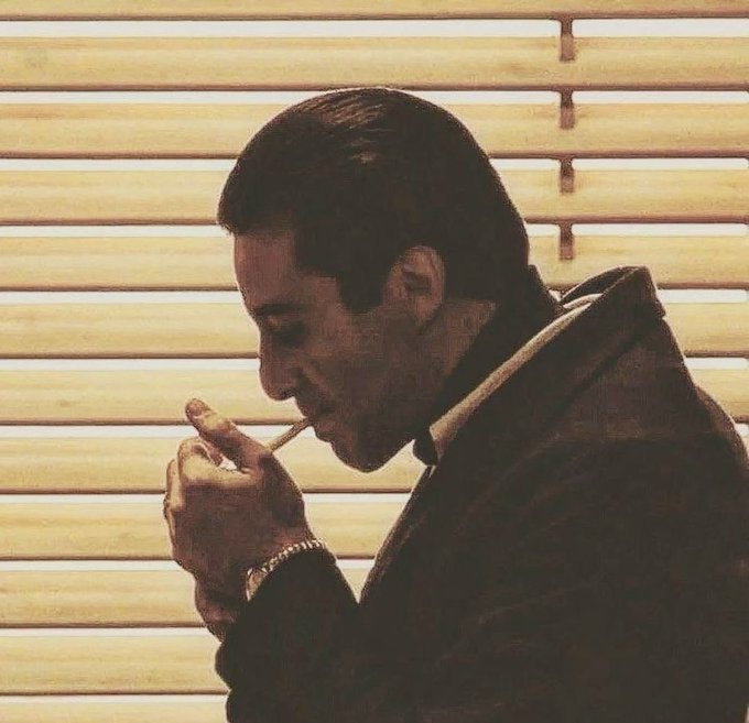 A vintage-styled photograph of a man in a dark suit lighting a match or cigarette, silhouetted against horizontal window blinds.