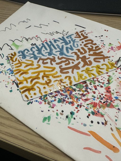 A white tile or canvas covered in colorful graffiti-style squiggles (blue, brown, yellow) sits on top of a splattered paint artwork on a wooden desk.