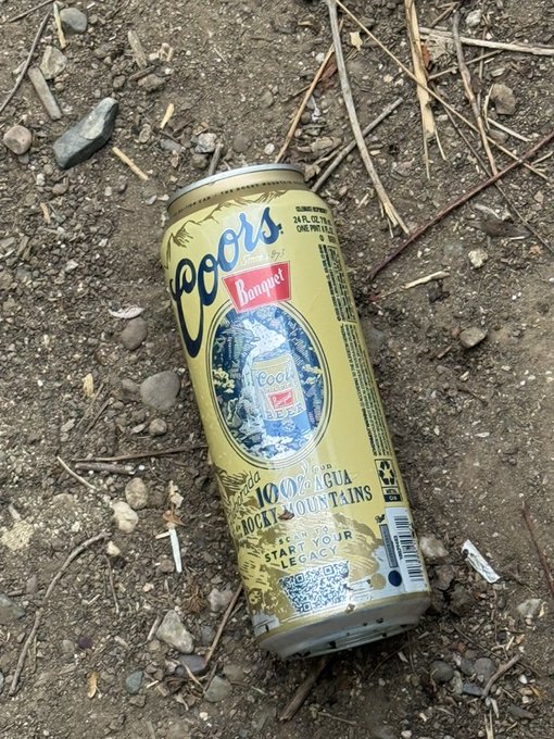 A discarded Coors Banquet beer can lies on rocky dirt ground, with the label reading '100% AGUA DE LAS MONTAÑAS' and a QR code visible.