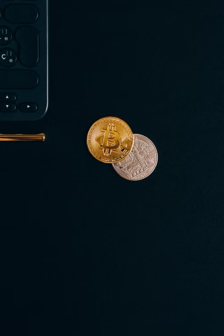 Two physical Bitcoin coins (one gold, one silver) lie on a dark surface next to a black calculator and a gold pen.