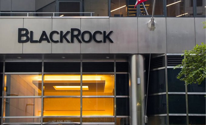 The exterior entrance of a BlackRock office building, showing the company name in large black lettering above glass doors with warm yellow lighting inside.