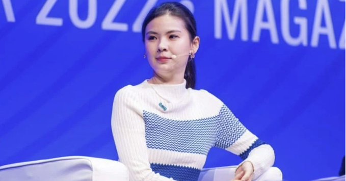A young woman with dark hair wearing a white and blue patterned sweater speaks at a panel, with partial text '2020' and 'MAGA' visible on the blue backdrop.