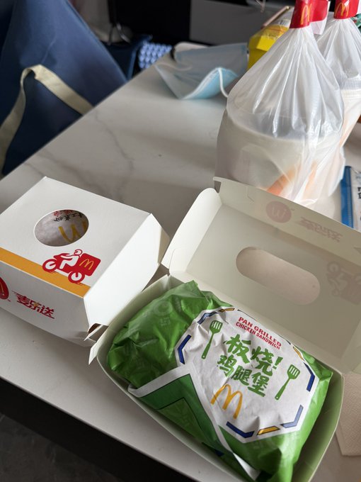A McDonald's meal sits on a tray table, featuring a green-wrapped "板烧鸡腿堡" (Grilled Chicken Burger) and a "麦乐送" (McDelivery) box, likely eaten during a train or plane journey, matching the post's claim of paying for a McDonald's meal with Bitget Wallet.