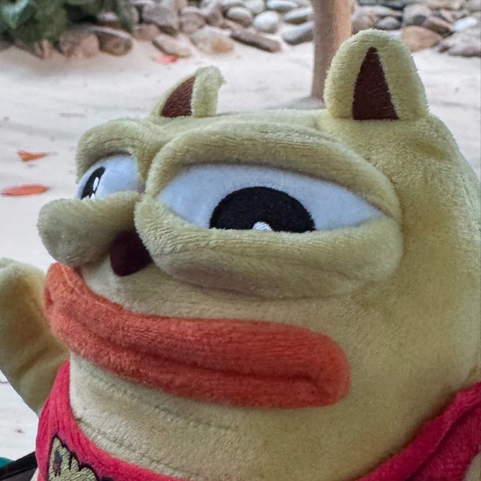 A close-up of a plush toy with a yellowish-green face, large googly eyes, an orange felt mouth, and a red shirt.