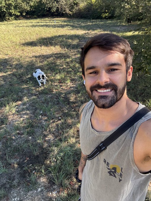 A bearded man in a beige tank top takes a selfie outdoors while a black and white dog runs toward him on a grassy path.