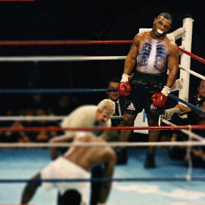 A vintage boxing match scene where a muscular Black boxer wearing red gloves stands in the ring looking down at his fallen opponent, while a referee and cornermen observe from the background.