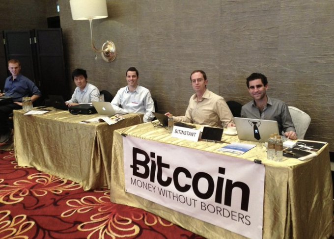 Five men sit behind a table draped in a gold cloth with a large banner reading "Bitcoin - MONEY WITHOUT BORDERS," along with a smaller "BITINSTANT" sign, all working on laptops in what appears to be a hotel conference room. This image captures an early Bitcoin industry gathering celebrating the cryptocurrency's first halving event, when BTC was valued at just $12—now legendary figures from the nascent crypto community.