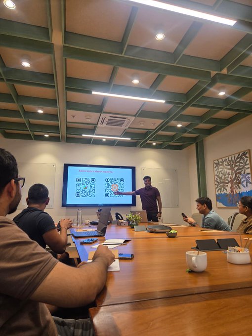 A presenter in a maroon shirt points at a screen displaying QR codes during a meeting in a modern conference room with attendees around a wooden table.