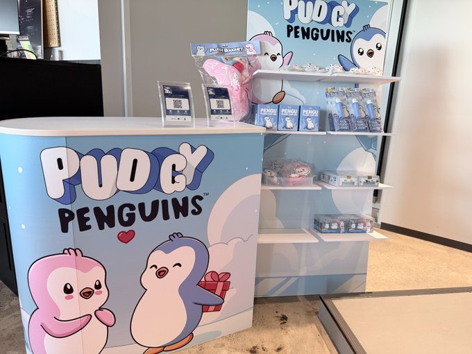 A merchandise booth displaying plush toys and packaged items for 'Pudgy Penguins' at what appears to be a convention or store.