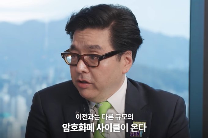 A man with dark hair and black glasses wearing a dark suit and green tie speaks in front of a window showing a city skyline, with Korean subtitles reading '이찬우는 다른 규모의 암호화폐 사이클이 온다'.