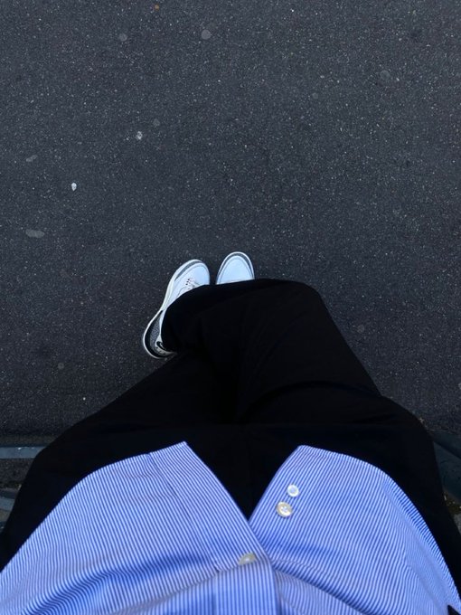 A first-person view looking down at white sneakers, black pants, and a blue striped shirt against a dark asphalt surface.