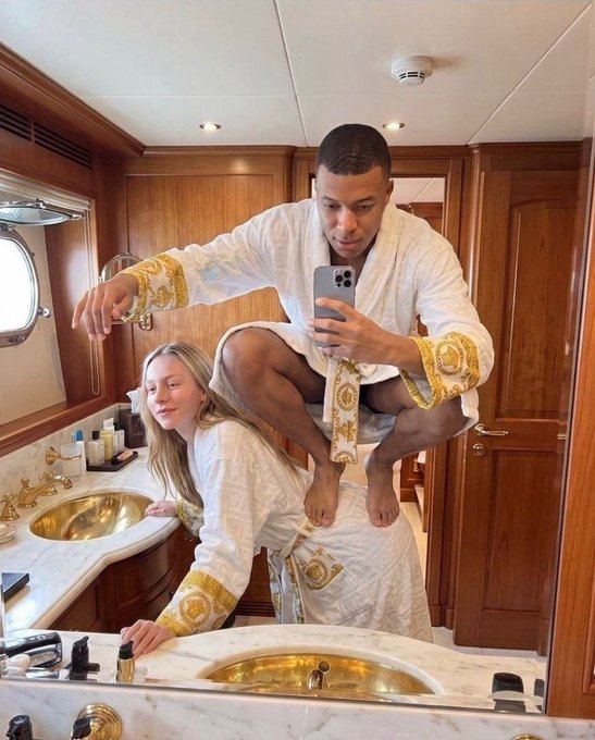 A man crouching on a woman's shoulders as she leans over a bathroom sink, both wearing matching white and gold Versace robes while taking a mirror selfie.