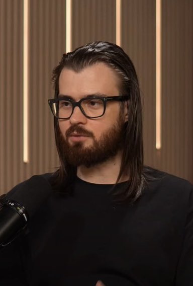 A portrait of a man with long dark hair, a full beard, and black-framed glasses, wearing a black shirt, sitting in front of a microphone against a paneled wall background with vertical light strips.