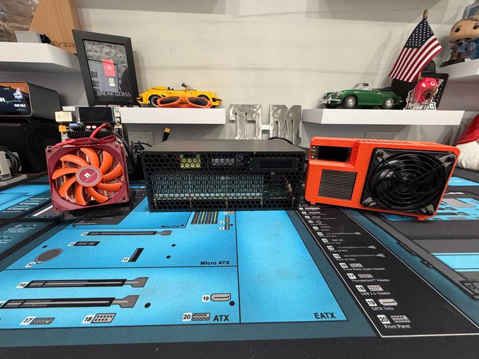 A collection of computer hardware components arranged on a blue motherboard schematic mat, including a red fan with a Thermaltake logo, a black server unit, and an orange speaker, with shelves displaying model cars and a 'BREAK THE RULES' sign in the background.