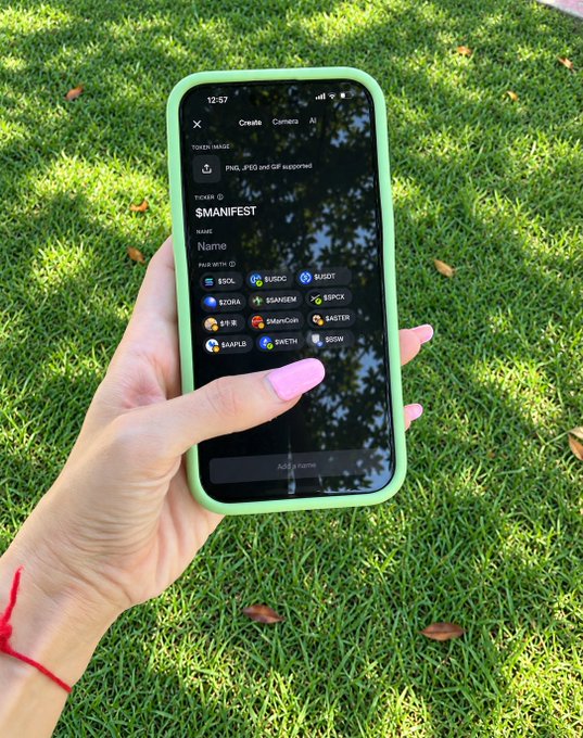 A hand with pink nails and a red string bracelet holds a smartphone with a green case, displaying a token creation app showing the ticker "$MANIFEST" and name "Name."