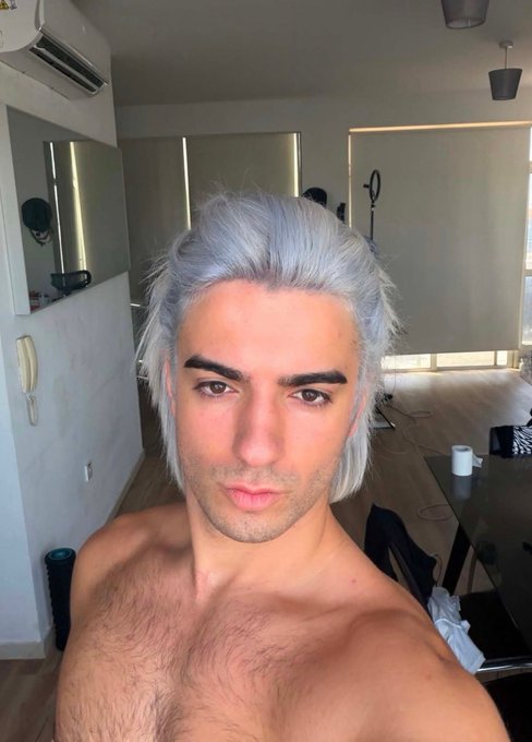 A shirtless young man with tousled silver/white hair takes a close-up selfie in what appears to be a studio or apartment interior.