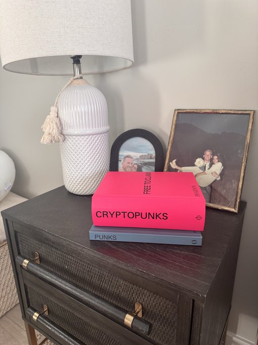 A home decor scene showing a dark wooden nightstand with a white ceramic lamp, framed wedding photos, and two stacked books labeled 'CRYPTOPUNKS' (pink) and 'PUNKS' (gray) from 'FACTUM', with a 'POW! POSTS' spine visible.