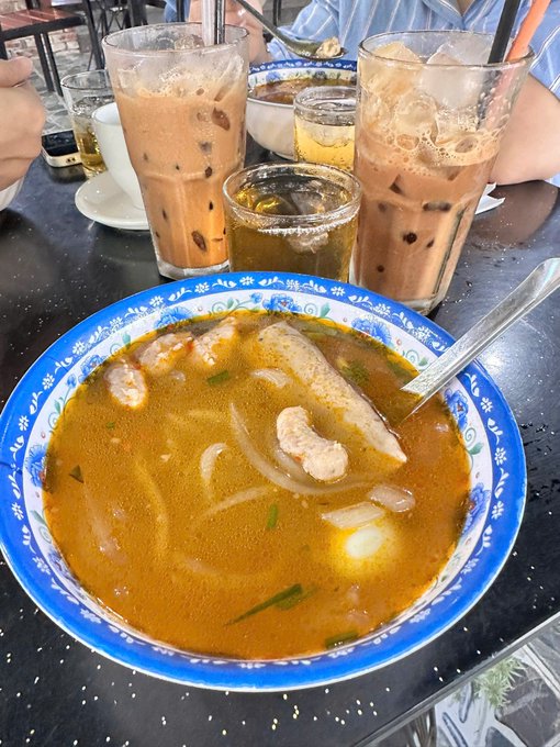 A bowl of shrimp soup with noodles and onions is served alongside glasses of iced tea and beer on a restaurant table.