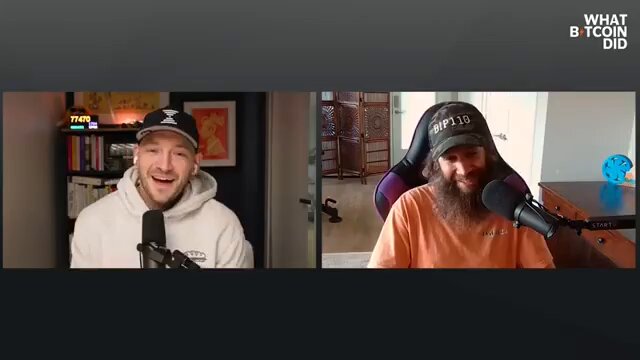 The image features two men sitting behind microphones, appearing to be engaged in an interview or discussion. One man has a beard and is wearing a white sweatshirt with a black logo, while the other man wears an orange shirt and also has a beard. The background of the image shows a bookshelf filled with various items such as books and a clock. The caption on the image reads "Why BIP-110 is a lot more likely to succeed than people realize." This suggests that the conversation might be related to cryptocurrency or blockchain technology, given their facial expressions and body language during the interview.