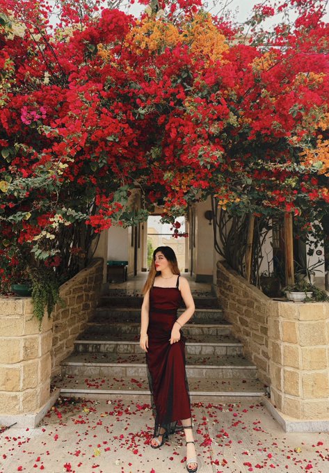 A woman in a long maroon dress stands at the base of stone steps beneath an archway covered in vibrant red and orange flowers.