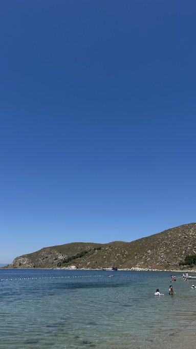 A serene beach scene unfolds under a vast, cloudless blue sky, with a rocky hillside in the distance and a few people wading and swimming in the calm, clear water—capturing the relaxed, optimistic vibe of a "good morning" with promises of a great day ahead.