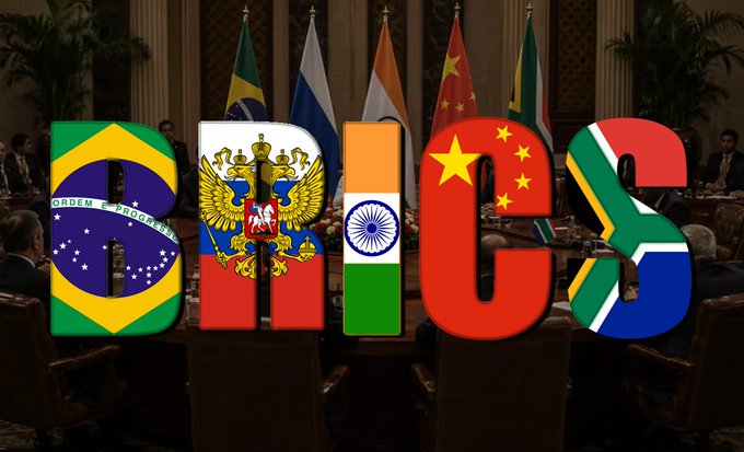 The letters "BRICS" are prominently displayed, each stylized with the flag of its respective member nation—Brazil, Russia, India, China, and South Africa—set against a backdrop of diplomatic flags and officials in formal attire, symbolizing the geopolitical bloc discussed in the post about its diminishing influence.