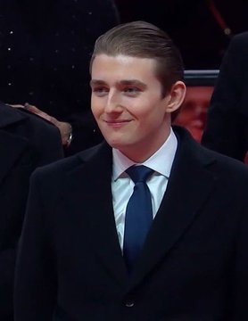 A young man with slicked-back brown hair wearing a dark suit, light blue shirt, and dark blue tie, smiling slightly at what appears to be a formal event.