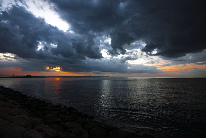 A moody sunset over a calm body of water, framed by dark, dramatic clouds and a rocky shoreline in the foreground.