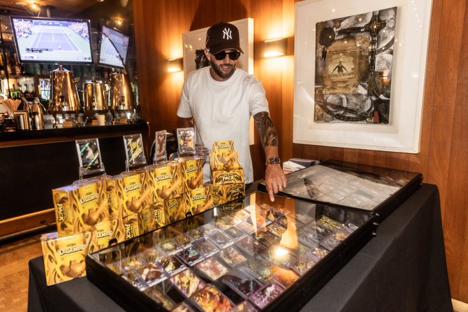 A man wearing a New York Yankees cap, sunglasses, and a white t-shirt stands behind a display of trading cards in cases and gold 'Packs' boxes.