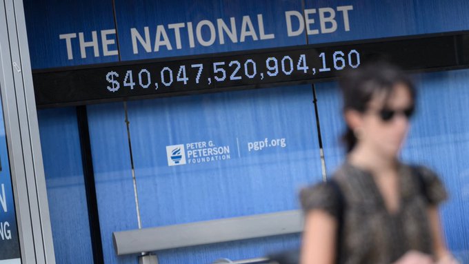A blurred woman walks past a digital "National Debt" display showing $40,047,520,904,160, sponsored by the Peter G. Peterson Foundation (pgpf.org), visually underscoring the post's warning that America's $40+ trillion debt crisis is driving the creation of an entirely new stablecoin-based financial system to replace the failing old one.